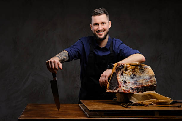 smiling chef cook holds a knife and posing near a table with exclusive jerky meat on dark background. - beef jerky meat smoked imagens e fotografias de stock