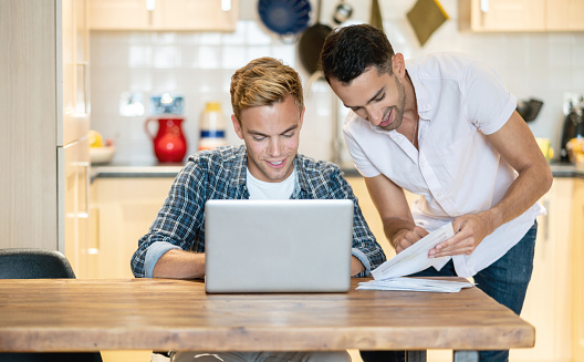 Happy gay couple paying bills online at home using a laptop computer and smiling â lifestyle concepts