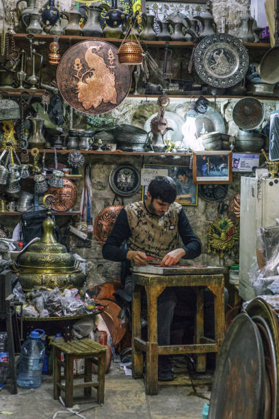 A local coppersmith is working in his little shop in a bazaar in Mardin, Turkey. Mardin is a city and multiple bishopric in southeastern Turkey. Copper working in Mardin is a traditional craft and dates back to several hundreds years ago.  Image has been captured from outside of the shop.