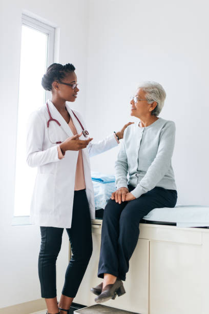 Senior female patient consulting with young female doctor A senior female patient sitting on a consulting bed and listening to a young female doctor standing next to her. hand on shoulder photos stock pictures, royalty-free photos & images