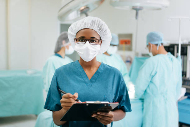 Female nurse holding clipboard in surgery room A young female nurse wearing protective uniform in the surgery room, holding a clipboard and looking at the camera with other healthcare workers in the background. surgical light stock pictures, royalty-free photos & images