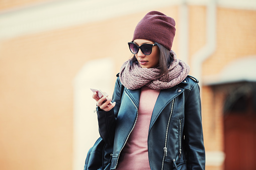 Young fashion woman in sunglasses using cell phone in city street  Stylish female model wearing black leather jacket beanie and knitted scarf outdoor