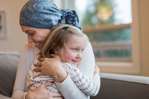 A beautiful young woman with cancer and wearing a headscarf holds her preschool-age daughter in her lap by their living window. She is squeezing her tight. Her daughter has a small smile and is hugging her mom affectionately.