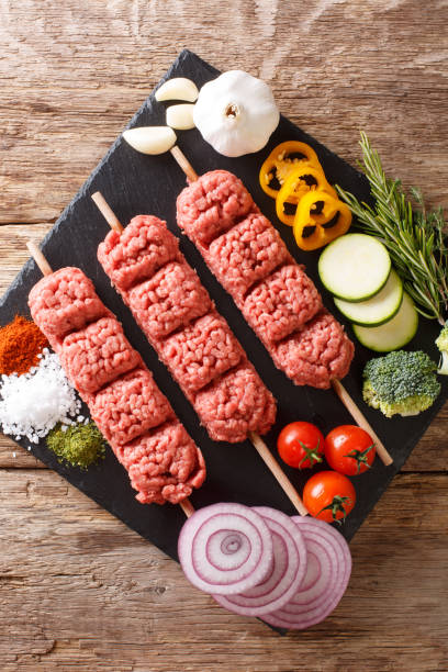 Homemade kebabs from raw minced meat on skewers with spices, herbs and vegetables close-up. Vertical top view