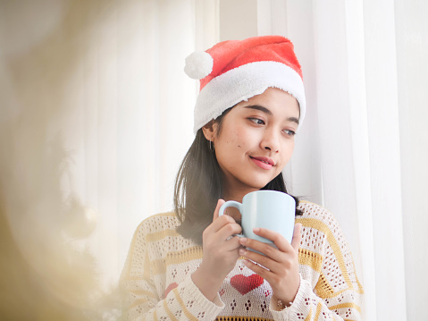 Asian woman wearing santa hat and holding coffee cup and look out window, lifestyle concept.
