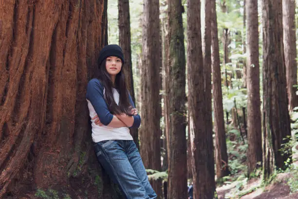 A candid portrait of a confident 11-year old eurasian girl standing against the trunk of a tree in the Redwood Forest, Rotorua