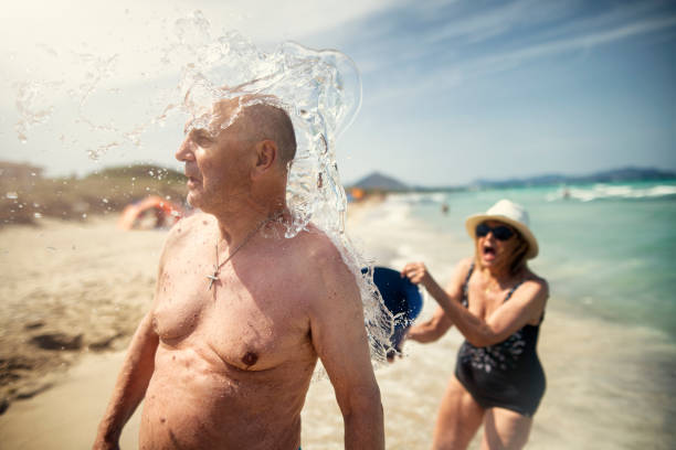 Senior woman surprising a senior man with a bucket of water on a beach. Senior couple having fun on a beach.\nNikon D850