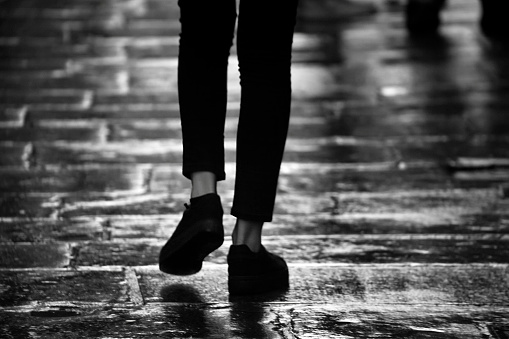 Young woman wearing jeans walking on wet street paving stone floor. Santiago de Compostela, Galicia, Spain. Black and white view.