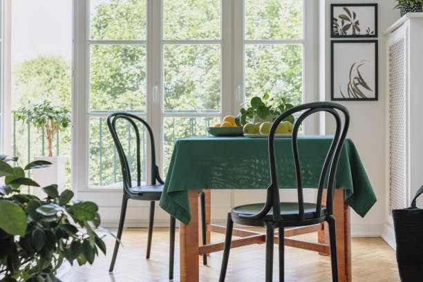istock Two black chairs standing by dining table with apples, lemons, fresh plant and green tablecloth in real photo of bright room interior with two posters and balcony with view 1065436730