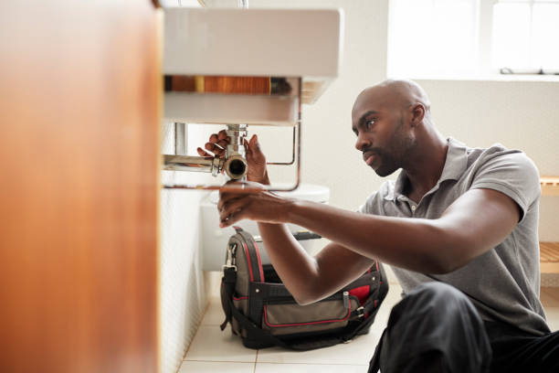 Young black male plumber sitting on the floor fixing a bathroom sink, seen from doorway Young black male plumber sitting on the floor fixing a bathroom sink, seen from doorway plumbing stock pictures, royalty-free photos & images