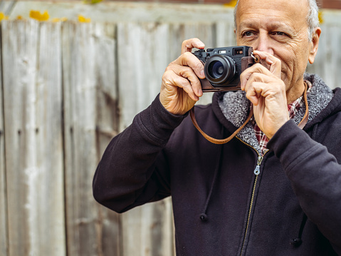 Senior man in fall outfit with retro digital camera standing in front of old wooden fence.