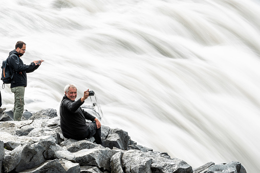 Dettifoss, Iceland - June 16, 2018: Largest waterfall in Europe closeup of happy man photographer with camera smiling, gray grey water, rocky cliff, people tourists on top, smooth long exposure water