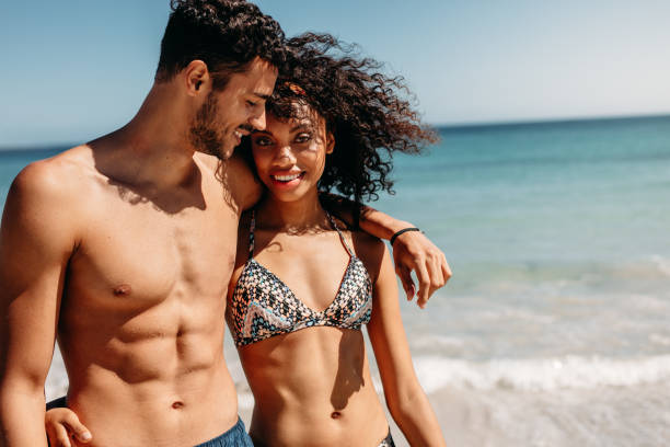 Couple walking on beach together Romantic couple walking on beach together on a sunny day. Smiling man walking on beach with girlfriend putting his arm around her shoulder. black woman bathing suit stock pictures, royalty-free photos & images