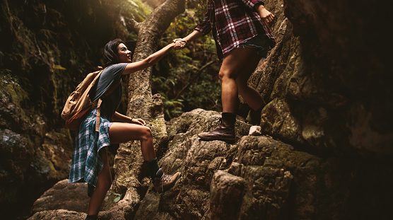 Woman holding hand of female friend while climbing a rock during mountain hike. Woman helping friend on a hike through extreme terrain in mountain.