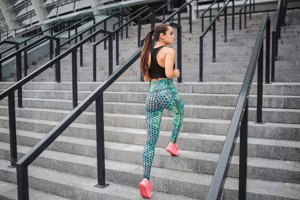 istock Model in good shape stands on steps and poses on camera. She shows her buttocks. Young woman wears sports uniform. She looks to right. Model stands there alone. 1065475448