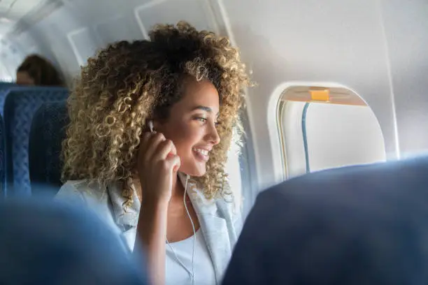 A young woman with curly hair sits in a window seat on a plane. She has her earbuds in as she looks out of the window and smiles.