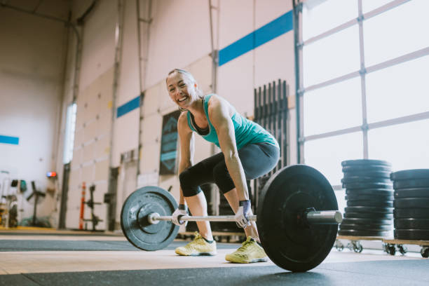 Mature Woman Practicing Deadlifts in Cross Training Gym A woman in her 50's enjoys weight training in a brightly lit gym, smiling at the camera. woman weight training stock pictures, royalty-free photos & images