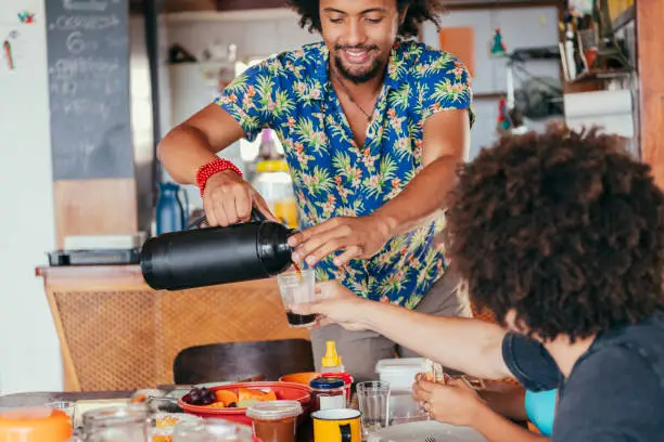 Man in Hawaiian shirt with Afro serving coffee to friend from flask, sitting at table in kitchen