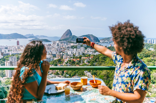 Brazilian man and woman sitting at table on vacation, eating breakfast on terrace, looking away, photographing Sugar Loaf Mountain