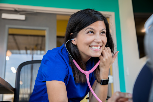 Female Asian nurse or doctor using a stethoscope while meeting with patient.  Doctor is wearing scrubs and sitting down.