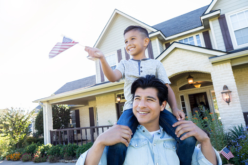 Cheerful mid adult Hispanic man carries his son on his shoulders. The boy is waving a small American flag.