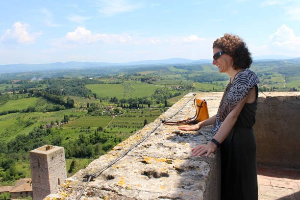 American woman traveller stands at the top of the tallest tower in San Gimignano, Italy looking over the city and Tuscan landscape. stock photo