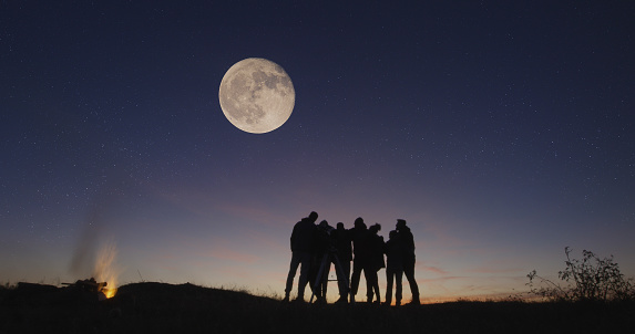 Silhouettes of people gathering together in nature looking on big moon with burning campfire near in night