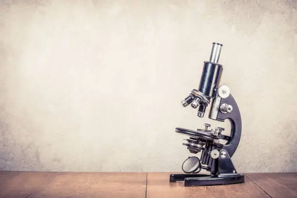 Photo of Retro old scientific laboratory microscope circa 40s on wooden table front concrete wall background. Vintage style filtered photo