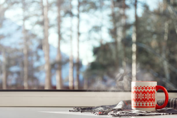 Steaming cup of hot drink on the window sill, with winter landscape in snow. Cozy home, winter weekend and holidays, Christmas morning concept.