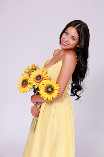 Young woman in tanned skin tone wearing summer fashion dress and holding a bunch of sunflowers in white isolated background