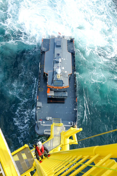 Manual high worker offshore climbing  down from wind-turbine on ladder and transfer vessel in waiting for him stock photo