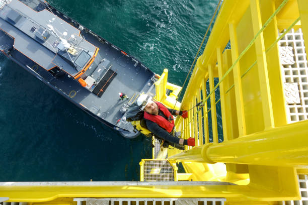 Manual high worker offshore climbing  down from wind-turbine on ladder and transfer vessel in waiting for him stock photo