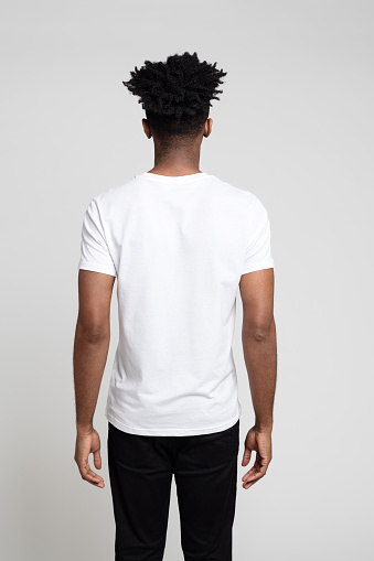 Rear view portrait of young afro american man standing facing gray background. Man in white t-shirt and black jeans.