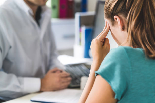 donna che soffre di mal di testa o emicrania. appuntamento con il medico in sala studio. signora malata e malata con stress, traumi o burnout. giovane paziente nel dolore che tiene la testa con le mani. - male doctor foto e immagini stock