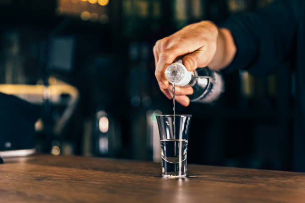Barmen hand with bottle pouring beverage into glass. Bartender pouring strong alcoholic drink into small glass on bar, shots, on bright background