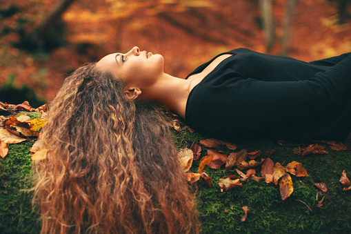 Beautiful young woman lying on tree trunk in forest and looking up. She has amazing long curly hair. Tree trunk is covered with moss and leaves colored with autumn.