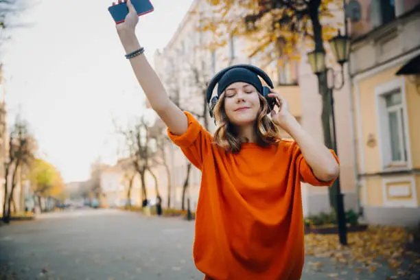 Photo of cute young girl listening music in headphones, dansing and holding mobilephone in hand, urban style, stylish hipster teen in black hat listen music on autumn city street, orange crazy street style