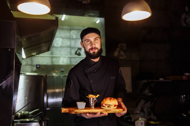 Smiling master chef in uniform looking at camera, holding plate with served burger