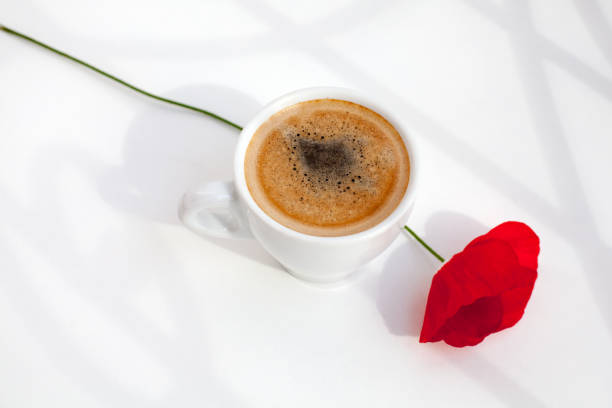 White mug of hot black coffee with foam and red poppy flower with green stem on white table in morning sunlight and beautiful shadows top view