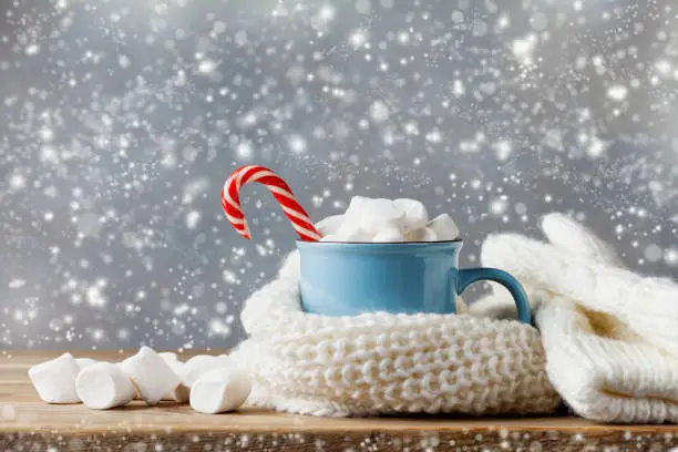 Winter lifestyle with cup of hot cocoa with marshmallows and knitted scarf on wooden background. Snow effect.