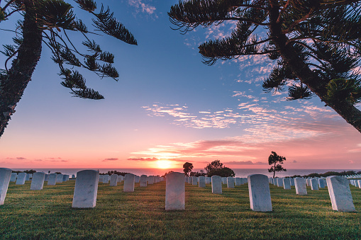 Fort Rosecrans National Cemetery, Point Loma, San Diego, California, USA.  Monument headstones along the coast during a pink, orange, and blue sunset with clouds and the ocean in the background and trees, grass, and tombstones in the foreground.