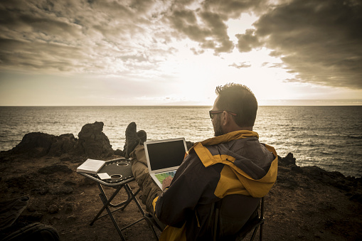 man traveler working during the sunset on a cliff with the ocean and the sun in front of him, freedom from office doing business everywhere like a digital nomad enjoying freedom and discovering the world