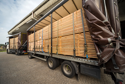 Semi-truck and trailer with stacked wooden planks in warehouse for wood and timber construction material. Blue sky, outdoors.