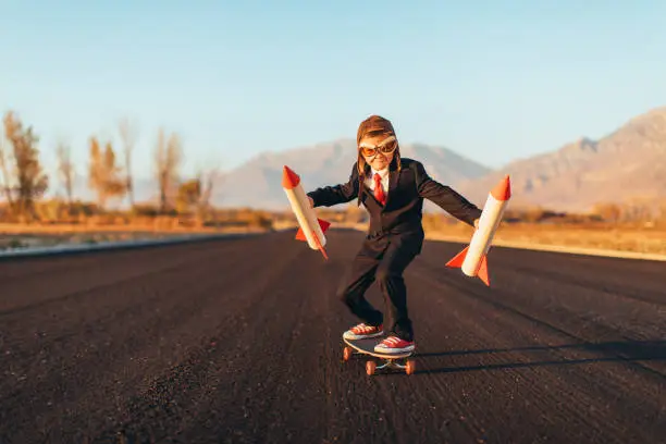 A young business boy dressed in a business suit, aviator cap, and goggles, holds rockets while standing on a skateboard. He is ready to launch his startup business the way forward. This entrepreneur is eager to make money with his new ideas. Image taken in Utah, USA.