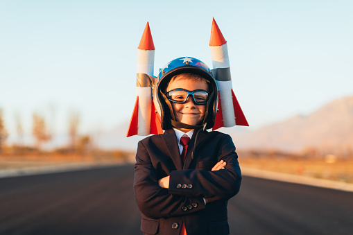 A young business boy dressed in a business suit stands with arms folded and rockets strapped to his helmet. He is ready to launch his startup business to the sky. This entrepreneur is eager to make money with his new ideas. Image taken in Utah, USA.