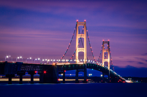 The tall towers of Michigan's majestic Mackinac Bridge shine brightly as the evening sky glows with shades of purple during twilight. This time lapse image features streaks of light from a large ore carrier cruising beneath the span as motorists cross over the Straits of Mackinac on the roadway above. This engineering marvel connects Michigan's upper and lower peninsulas which are nearly five miles or 8 Km apart. Impressive architecture background with copy space.