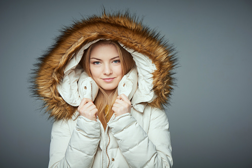 Portrait of beautiful girl in winter coat smiling looking at camera muffling in hood, on grey studio background