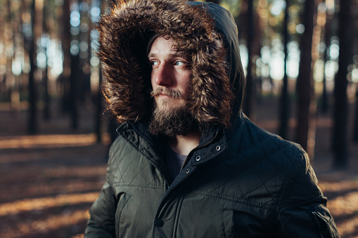 Portrait, close-up of young stylishly serious man with a beard dressed in rgreen winter jacket with a hood and fur on his head stands against the background of pine forest