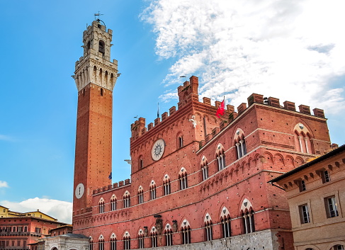 A suggestive view from below of the majestic Torre del Mangia (Mangia tower) and the Palazzo Pubblico (Public Building) in the Piazza del Campo, in the medieval heart of Siena, in Tuscany. Built in 1290 as the seat of the Council of Nine and of the executive power of the medieval city, the majestic Palazzo Pubblico is still the seat of the municipal government of Siena. The Torre del Mangia was built between 1338 and 1348 on a project by Giovanni Balduccio, known as the 'Mangia', becoming the symbol of the political and military power of Siena compared to its rival Florence. Center of the city life since 1169, Piazza del Campo or simply Campo is one of the most beautiful and famous squares in the world for its particular shell shape. In this space every year the seventeen historical districts of Siena compete in the Palio, one of the oldest horse races in the world. Siena is one of the most beautiful Italian cities of art, in the heart of the Tuscan hills, visited for its immense artistic and historical heritage and for its famous popular traditions. Since 1995 the historic center of Siena has been declared a World Heritage Site by UNESCO. Image in high definition quality.