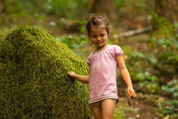 A cute little girl is sitting in nature under the tree on a summer day and looking at the camera.